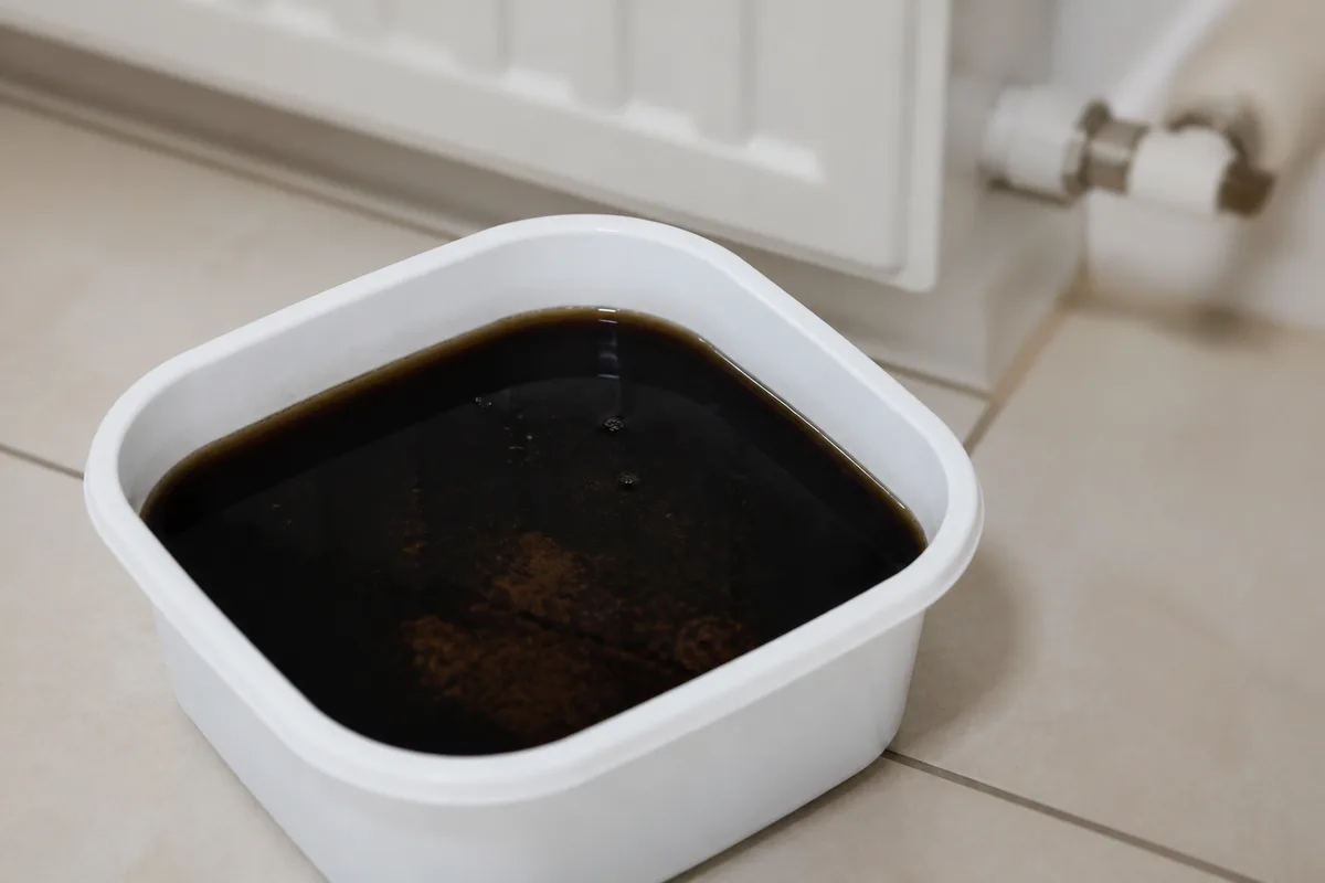 Récipient blanc rempli d&rsquo;eau de purge noire posé au sol devant un radiateur en fonte